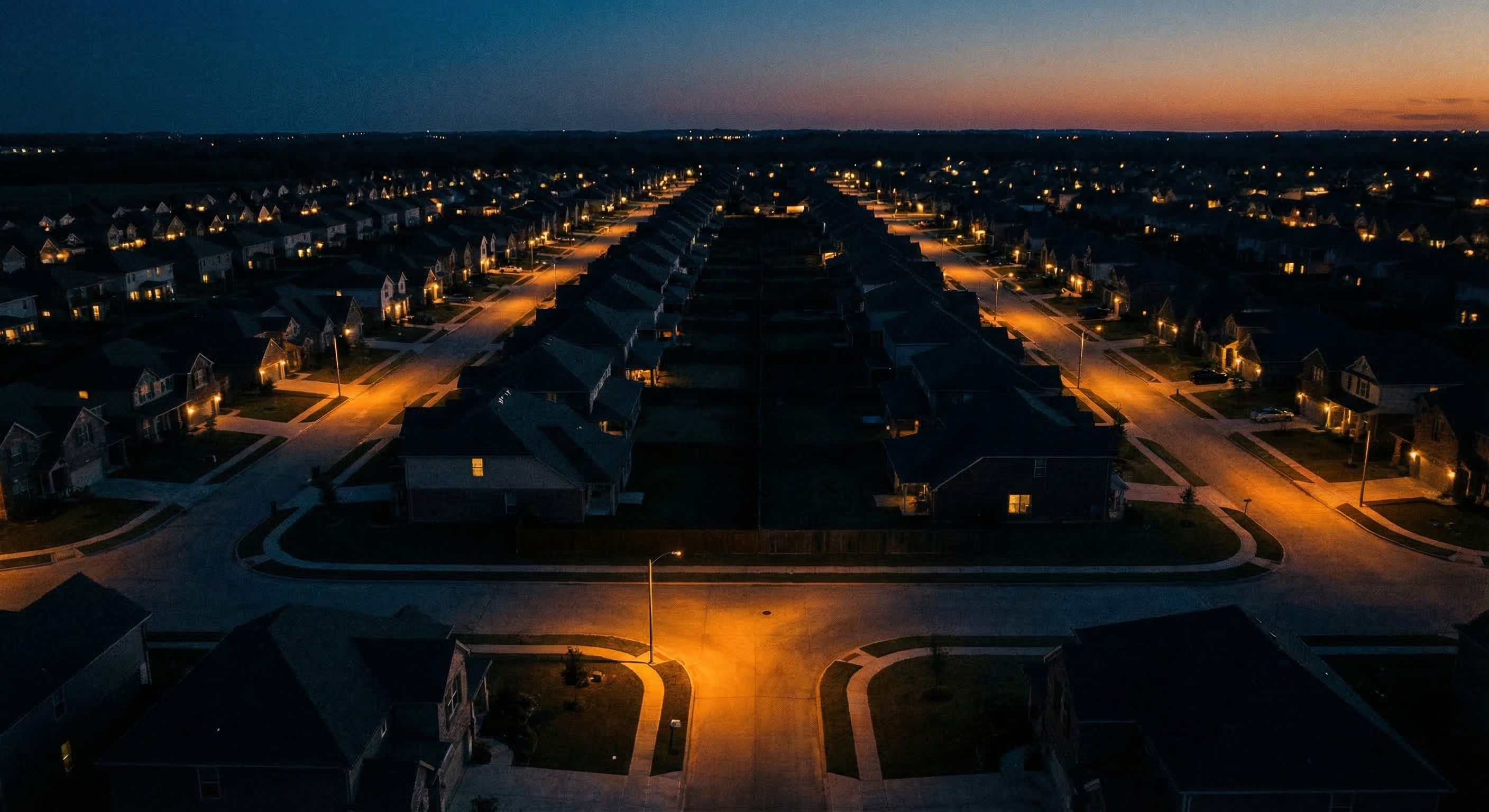 Aerial view of a still American suburban neighborhood at dusk, rows of houses with glowing windows under deep blue and amber light