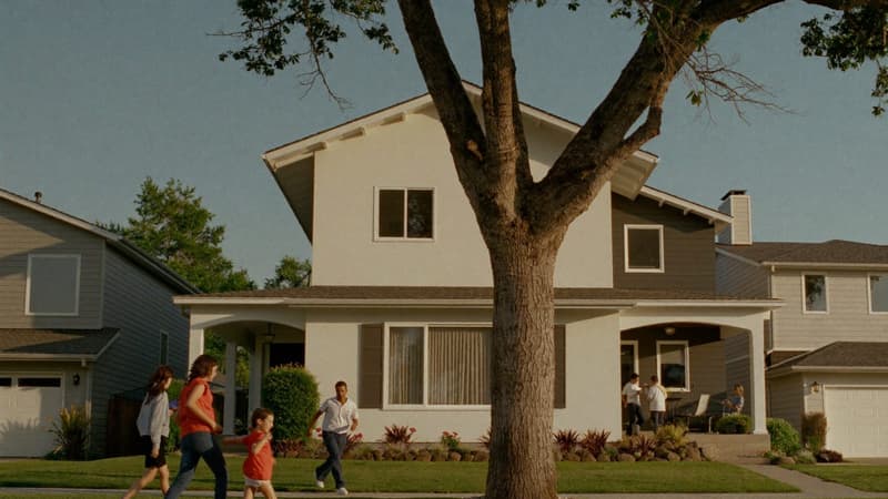 A warm American residential street at golden hour lined with mixed housing — single-family homes, a duplex, and a small apartment building — as a family walks toward a home for sale.