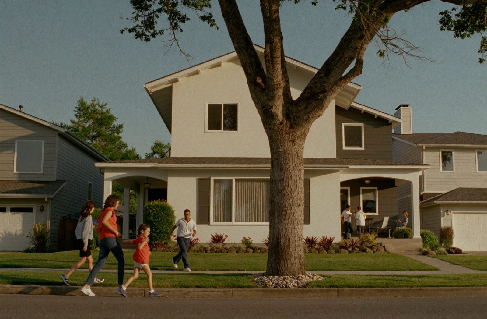 A warm American residential street at golden hour lined with mixed housing — single-family homes, a duplex, and a small apartment building — as a family walks toward a home for sale.