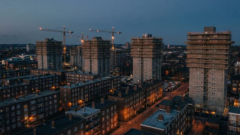 Two apartment towers in mid-construction at dusk, tower cranes still standing over their unfinished upper floors