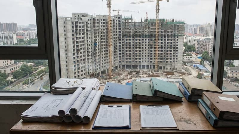 Affordable housing development complex under construction with stacked architectural cost estimate documents on a planning desk in a government office