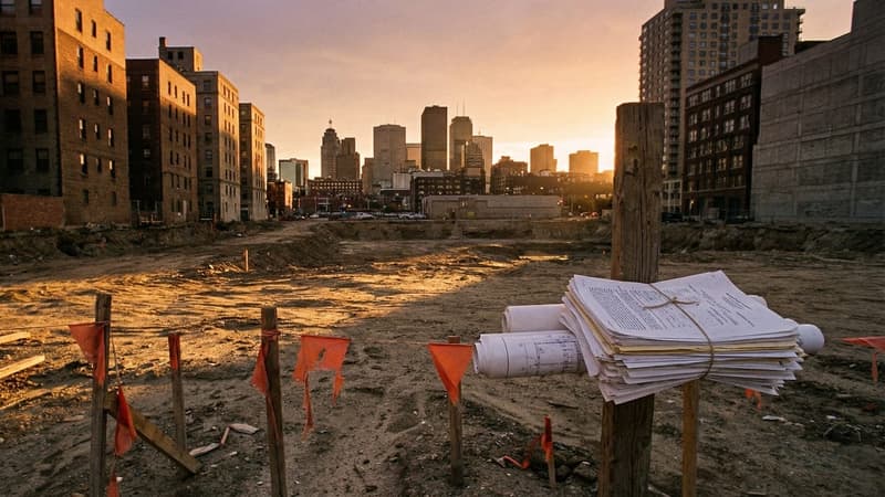 An empty urban development lot at golden hour surrounded by city buildings, with survey stakes and permit documents at the site boundary